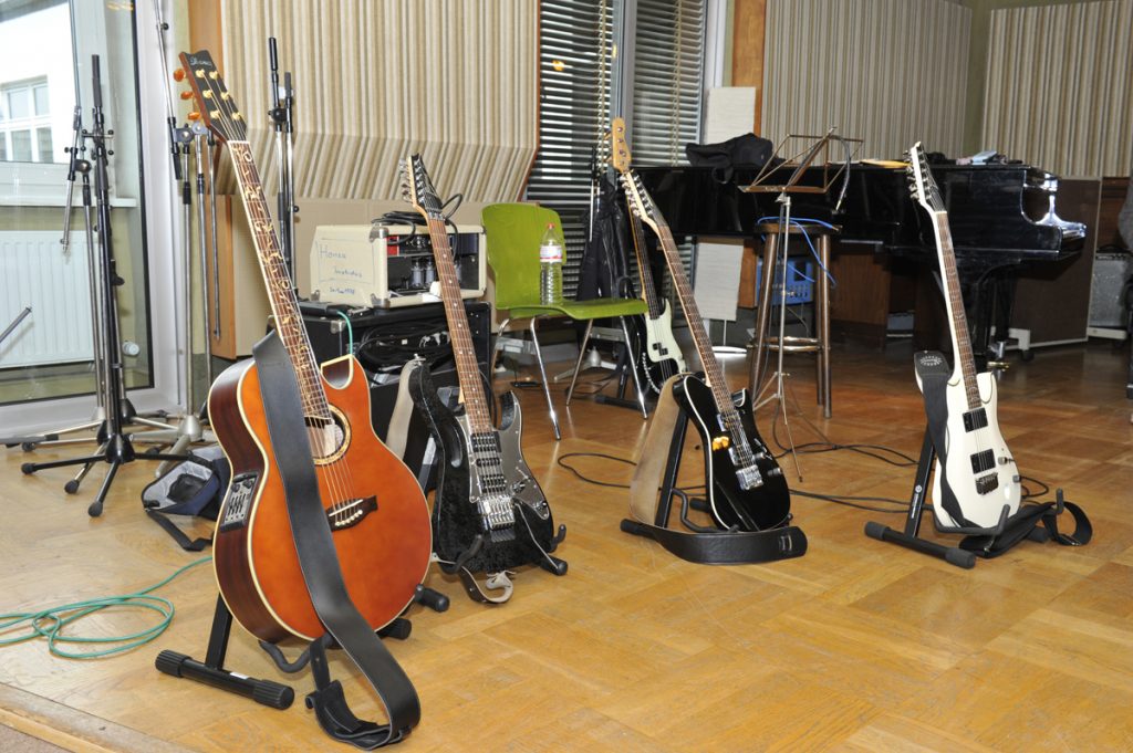 Akustikgitarre und drei E-Gitarren auf Ständern in einem Studio, dahinter ein Röhrenverstärker