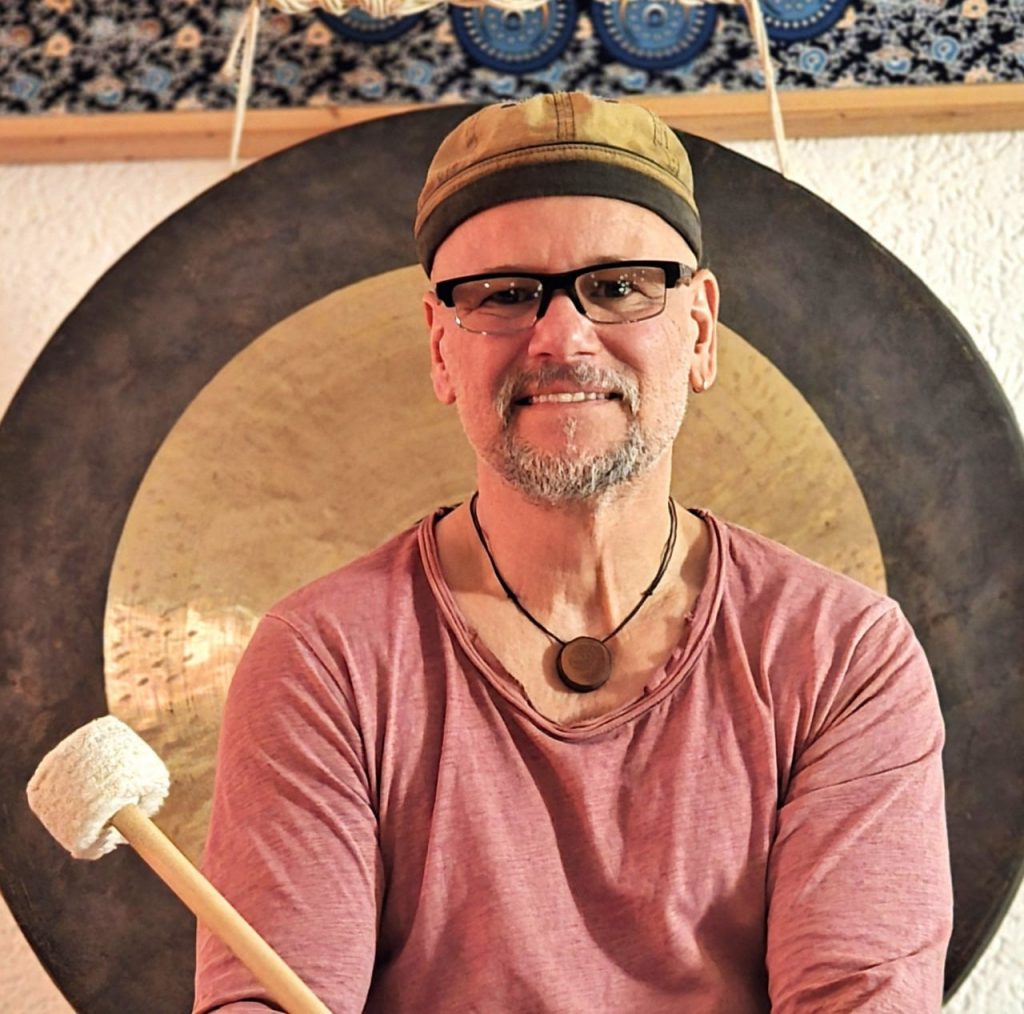 Joey Krüger steht lächelnd vor einem großen Gong und hält einen Gongschlägel in der Hand – Portraitbild für die Über-mich-Seite