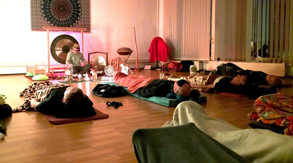 Live-Klangreise mit Joey: Teilnehmende liegen entspannt im Vordergrund, während im Hintergrund Gong, Klangschalen und weitere Instrumente gespielt werden